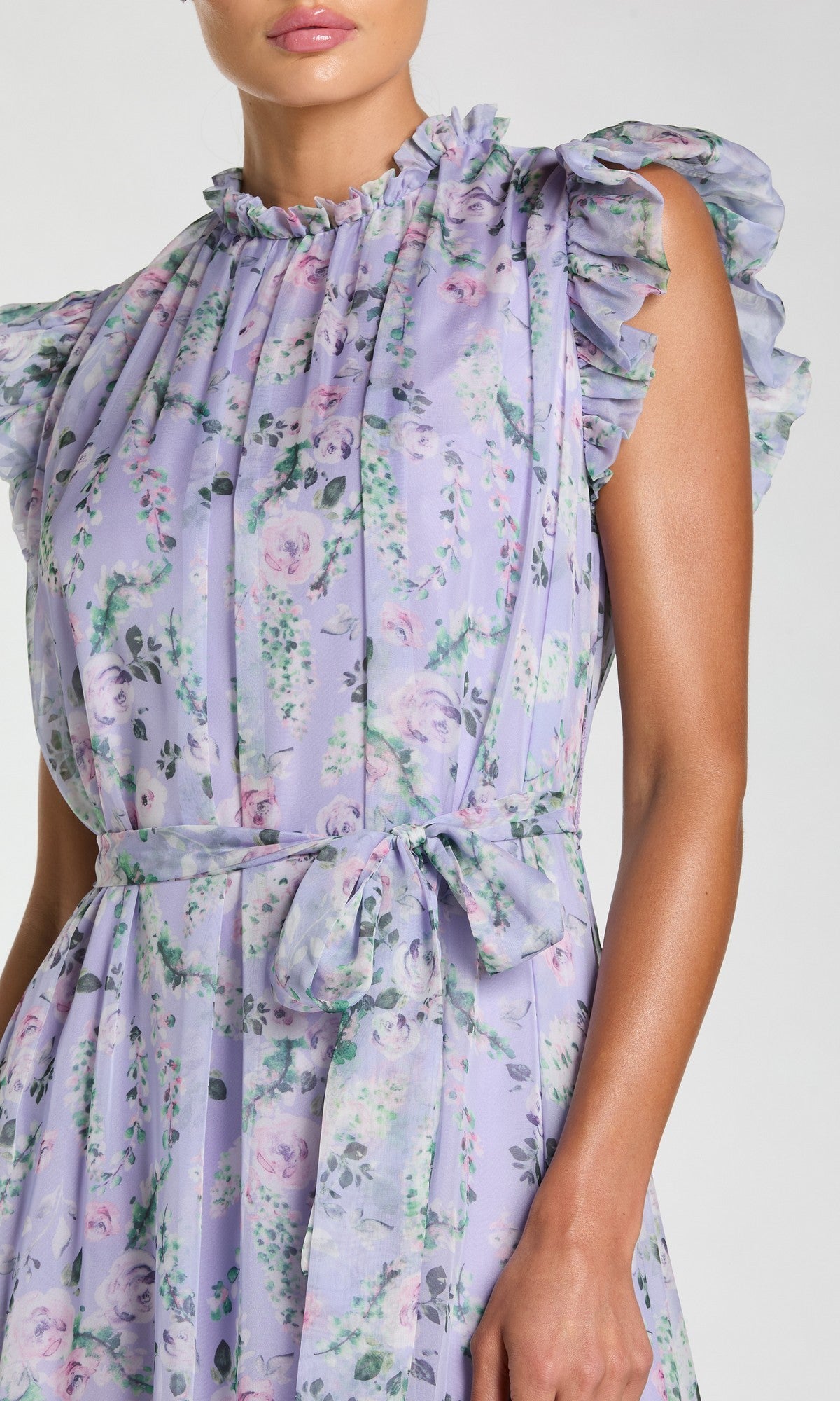 A woman wears the Mac Duggal 56116 Lilac Floral-Print Formal Gown, featuring a sleeveless design, high neckline, pleated details, ruffled accents, a tied waist belt, and an elegant floral print.